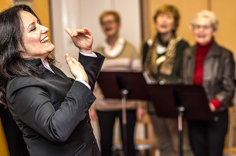 Fröhlich dirigierende Chorleiterin, im Hintergrund Teil eines Senior*innenchores