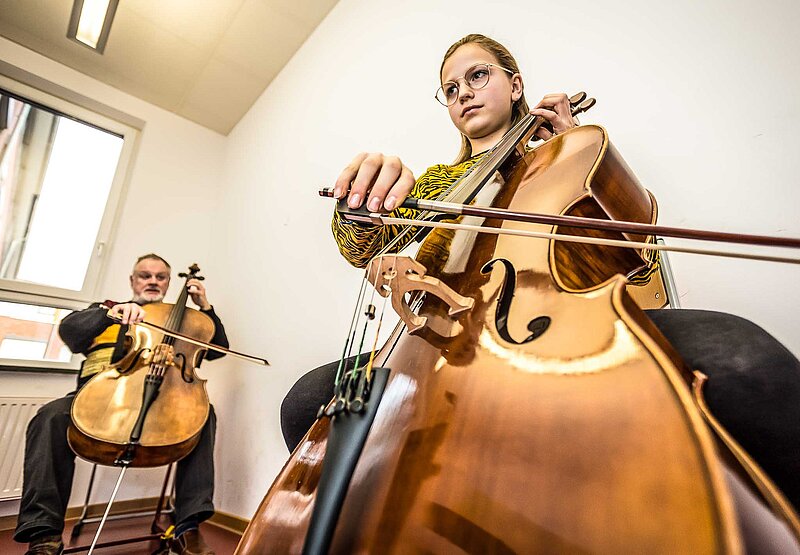 Jugendliche spielt konzentriert Cello, im Hintergrund der Cello spielende Lehrer; dynamische Weitwinkel-Froschperspektive