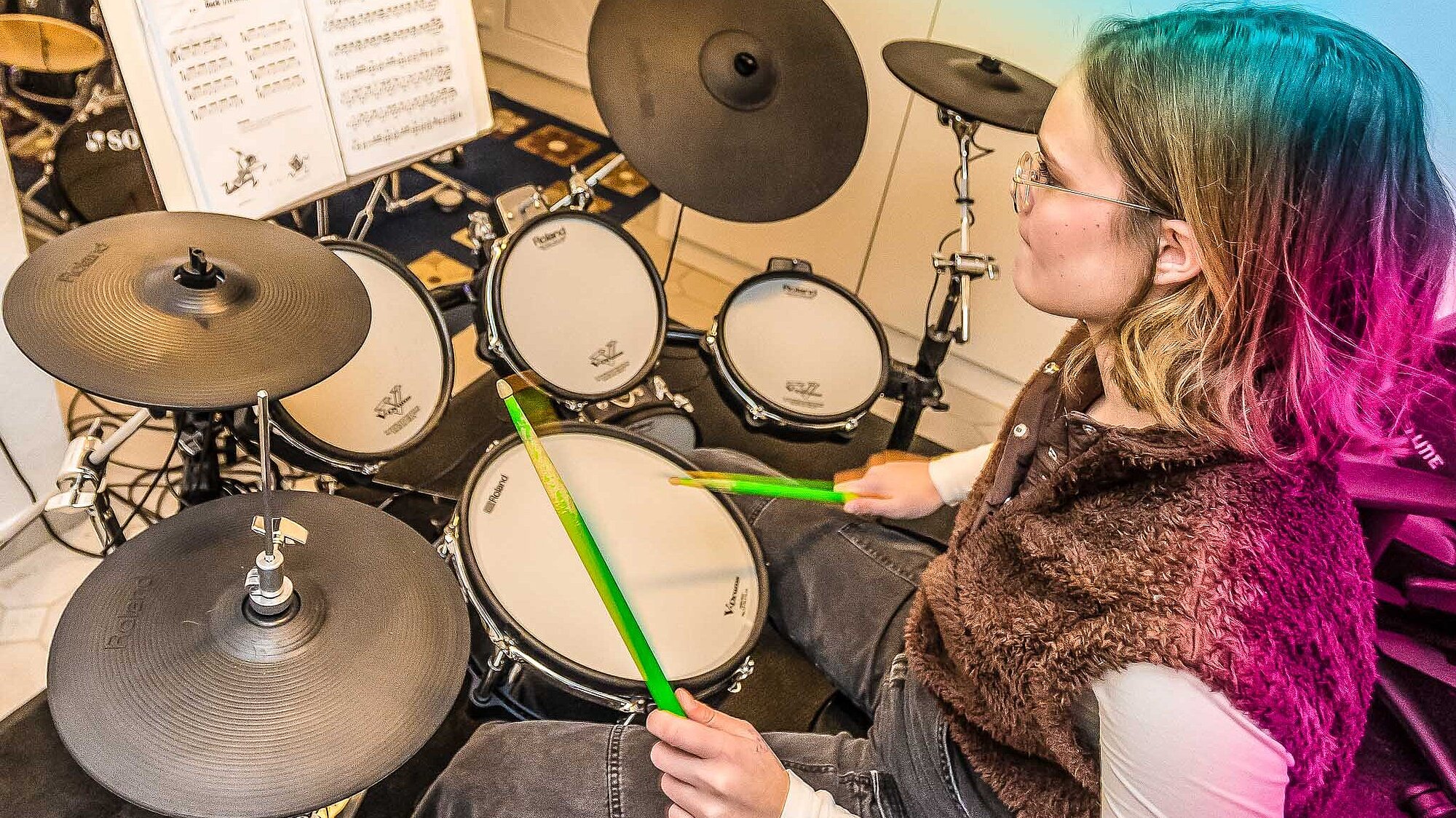Jugendliche übt konzentriert an einem E-Drumset; Im Hintergrund der Lehrer an einem eigenen Drumset