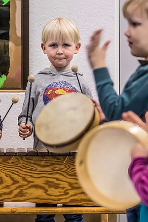 Zwei Vorschulkinder spielen auf Rahmentrommeln, ein weiteres schwingt im Hintergrund Schlegel für ein Xylophon